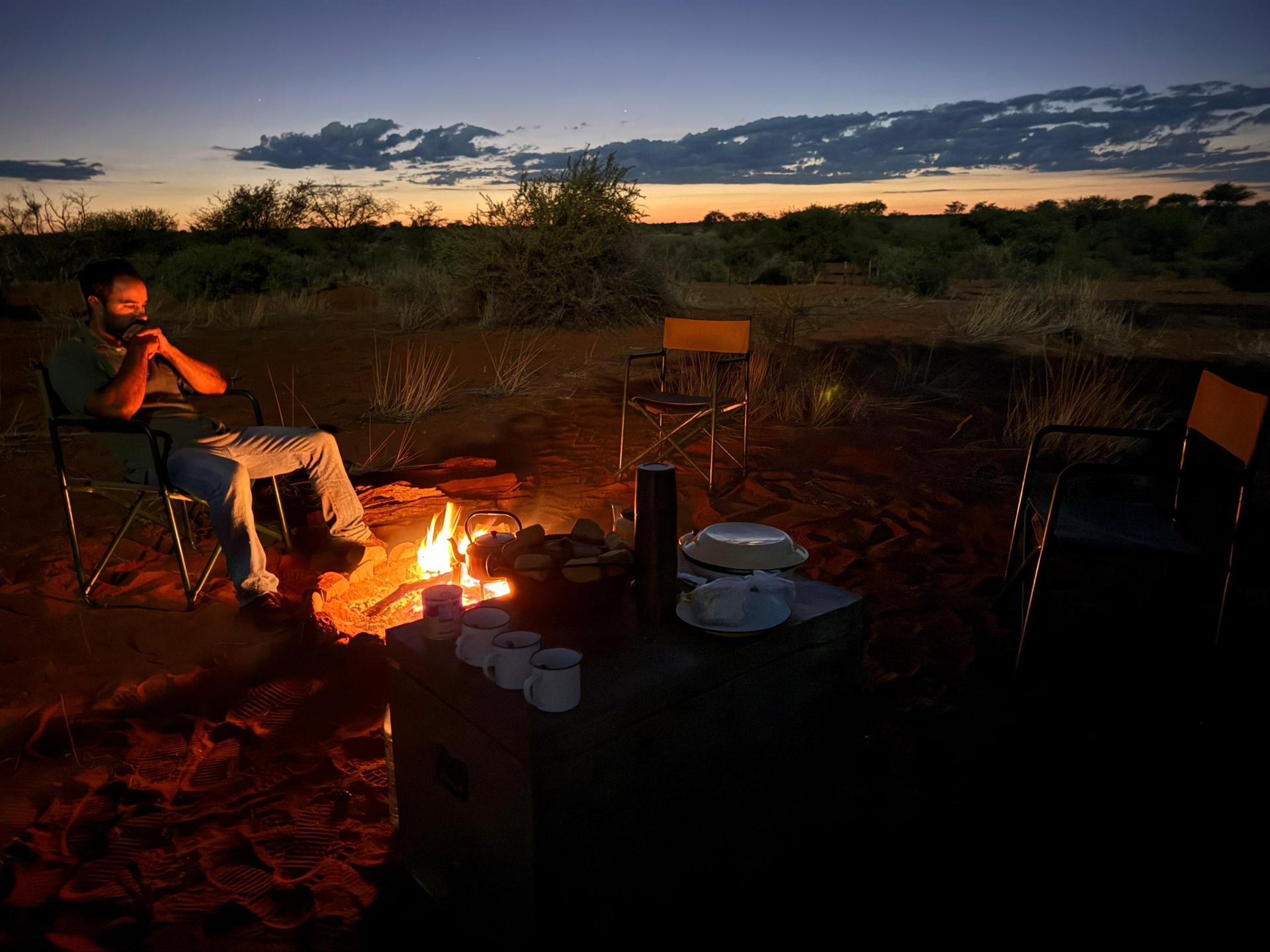 The camp fire glows against a wide Kalahari dusk